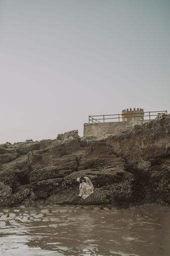 A young caucasian female with tattoos wearing a dress and straw hat sitting on the rocks