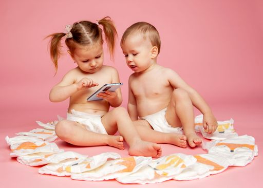 Two cute little babies sitting on the ground surrounded by pampers