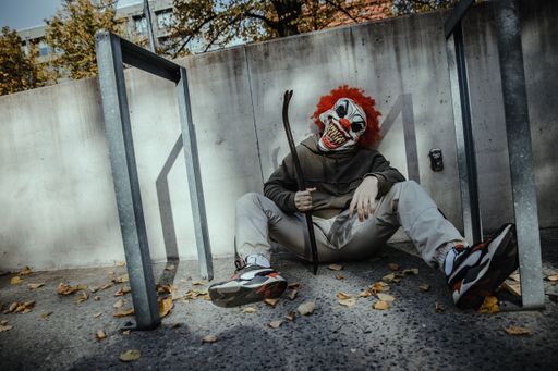 A young boy sitting in a corner outdoors with a scary mask of a clown