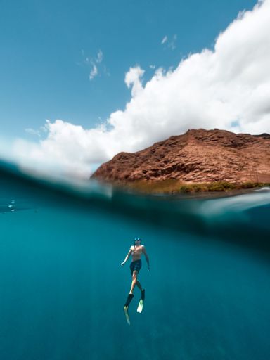 A mesmerizing shot of a male free diver swimming up to surface near to an island in bright sunlight against blue cloudy sky