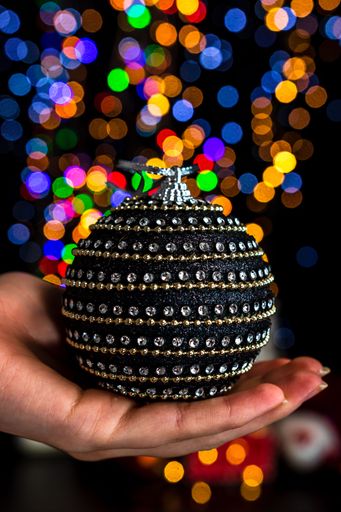 A vertical shot of a hand holding a Christmas black ball decoration with bokeh Christmas lights