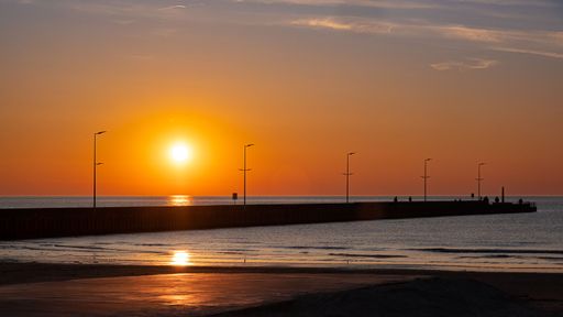 A tranquil scenery of a sunset reflecting on the ocean with silhouette of a pier at the coastline