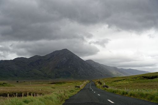 An empty road between green lands and mountains in the horizon with a cloudy gray foggy sky
