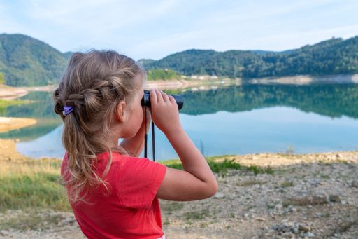 A closeup of a little girl looking through the binocular near a lake