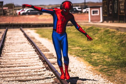 A closeup of person in Spider-Man costume wearing headphones with a backpack walking on sleepers on sunny day