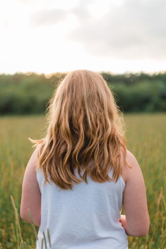 A back shot of a young blonde girl standing among long grass in field at sunset with blurred background