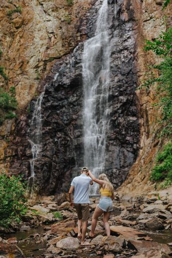 A backshot of a couple walking on rocks in daylight in fall brook falls against the waterfall in New Brunswick, Canada