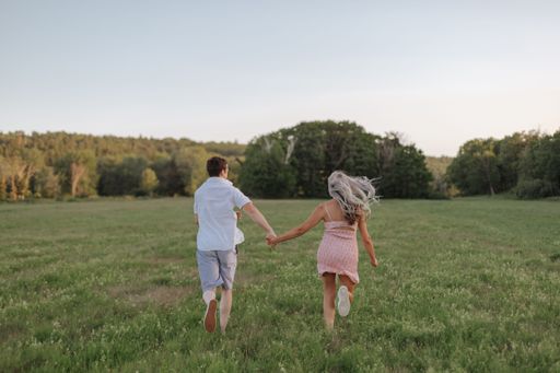 A backshot of a couple running on a green field on a sunny day against green trees, and the blue sky in New Brunswick, Canada