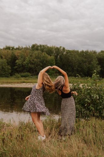 A vertical shot of two females dancing on tall green grass along the Miramichi River against green trees, and the cloudy sky in New Brunswick, Canada
