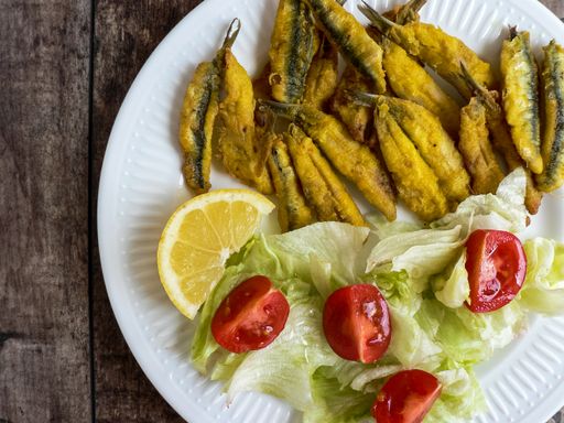 A closeup shot of fried anchovies with slices of tomatoes, lettuce and lemon on a white plate