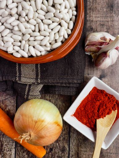 An overhead shot of an onion. garlic, carrot, paprika seasoning in a bowl, and white beans on a dark brown cloth placed on a wooden surface