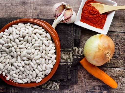 An overhead shot of an onion. garlic, carrot, paprika seasoning in a bowl, and white beans on a dark brown cloth placed on a wooden surface