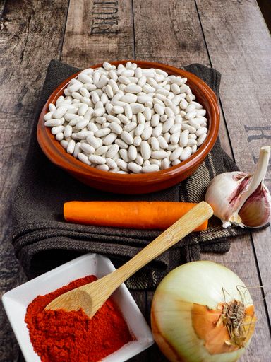 A high angle shot of an onion. garlic, carrot, paprika seasoning in a bowl, and white beans on a dark brown cloth placed on a wooden surface