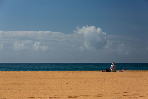 A scenic view of an adult man sitting on a sandy beach against a blue sea on a sunny day