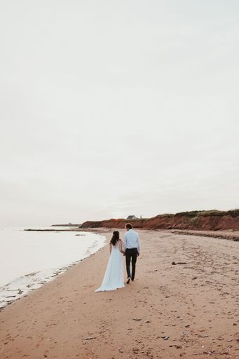 A vertical shot of a bride and a groom walking on the sand beach under a clear cloudless sky