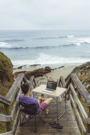 A vertical high angle shot of a young male sitting in front of a table with a laptop on the wooden staircase leading to the beach