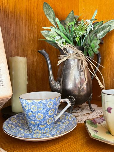 A vertical shot of an antique floral porcelain coffee cup with a vintage metal teapot in the background