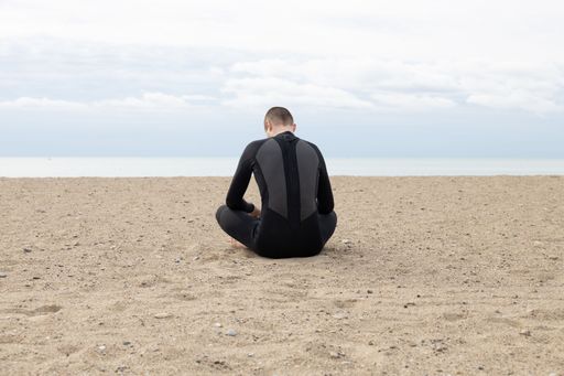 A Caucasian male in a scuba suit seen from behind, sitting on a sandy beach against a sea on a cloudy day