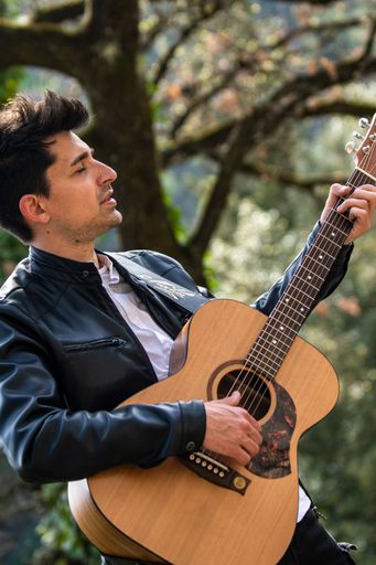 A vertical shot of a male in a leather jacket playing acoustic guitar outdoors