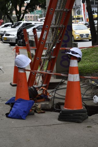 A vertical shot of a white hard hat on top of a safety cone with an extension ladder on a sidewalk