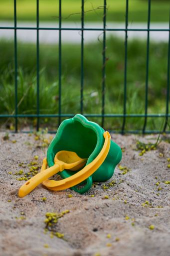 Vertical shot of a green bucket with yellow handle and a yellow shovel beach toys on sand and metal fence in the background
