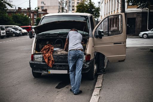 A man standing in front of the car hood on the street trying to repair it