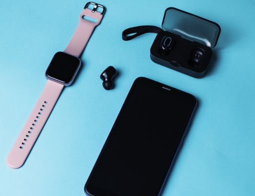 An overhead shot of smart phone,  smart watch with pink band, a wireless earphones case with one earphone  next to it on neon blue background