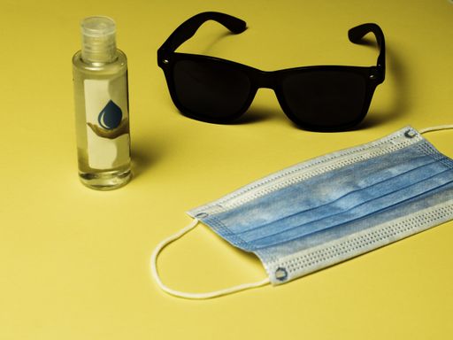 A medical mask, a bottle of hand sanitizer and black sunglasses laying on a solid yellow surface
