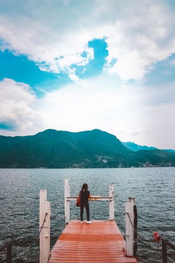 A vertical shot of a female standing on the dock near the sea enjoying the breathtaking view of the sea and rocky mountains
