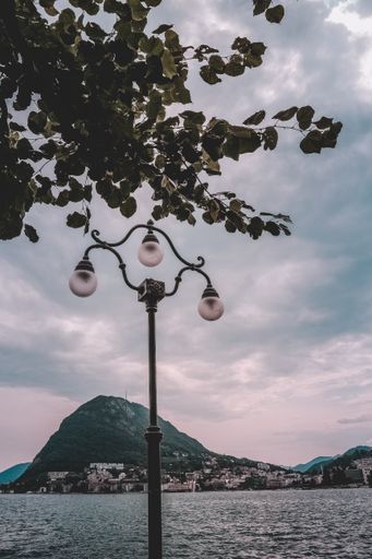 A vertical shot of a light pole near the wavy sea with the background of beautiful green mountains under the breathtaking cloudy sky