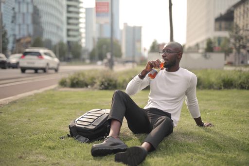 A stylish African male with glasses sitting on the grass and drinking juice under the sunlight