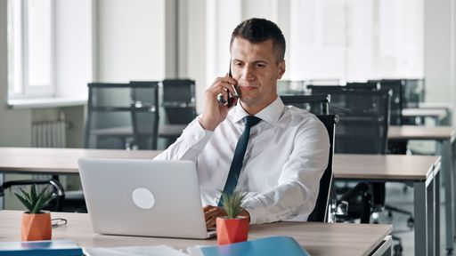 A man in a shirt and tie is sitting in the office in front of a laptop and talking on the phone