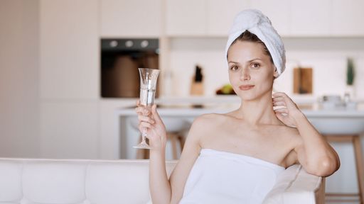 A woman wrapped in a towel after a shower sits on a sofa with a glass of champagne in her hand