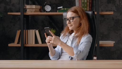 A red-haired white woman sitting in the office in front of a table and looking at phone