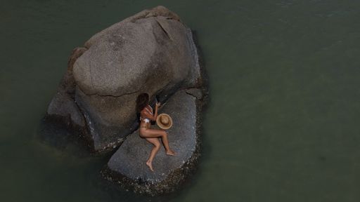 A top shot of a female wearing a Bikini laying down on a black rock on a sunny day in the middle of the water
