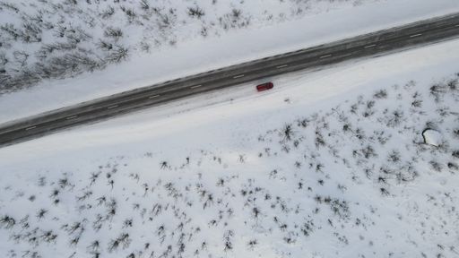 An aerial view of a vehicle on an asphalt road cutting through fields covered with snow