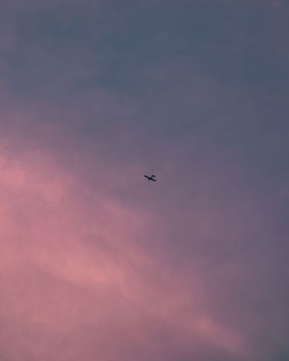 A vertical distant view of a jet flying away with cloudy sky in the background during sunset
