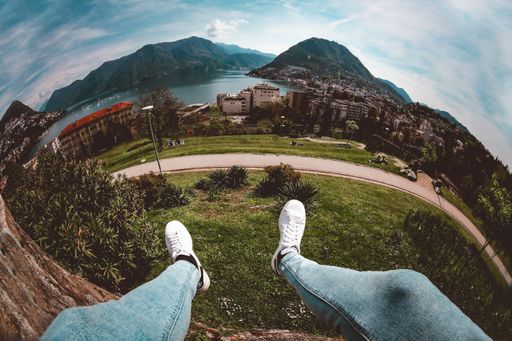 A high angle fish-eye shot of a person sitting on a cliff enjoying the breathtaking view of high buildings and green mountains near the sea