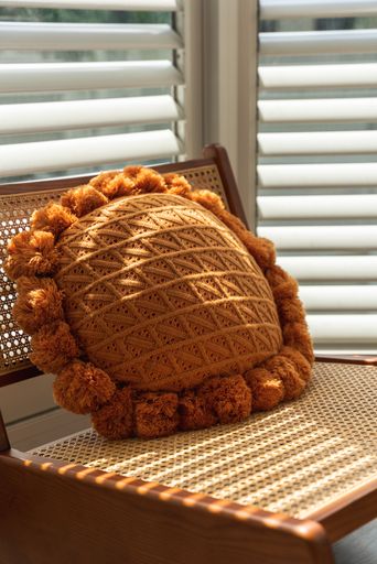 A vertical shot of a brown handmade cushion on a boho style chair in a room with horizontal blinds
