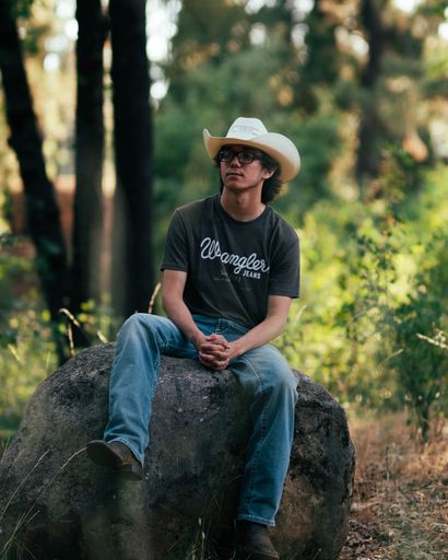 A man with a white cowboy hat and glasses sitting on a rock with a view of nature on the background