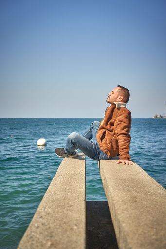 A vertical shot of a young Caucasian male sitting at the edge of a stone with Lake Geneva in the background, Switzerland