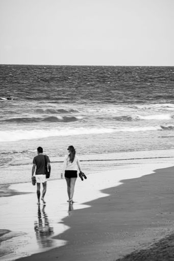 A vertical shot of a romantic couple walking on the sandy beach towards the ocean waves. Grayscale