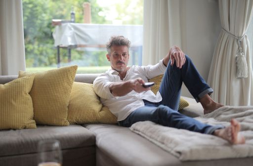 An adult male with a white shirt lying on the sofa and holding a remote control under the sunlight through the windows