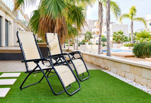 The two lounge chairs on artificial grass with modern apartments and a swimming pool in the background