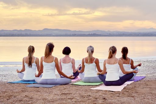 A Back view of six Caucasian women group yogi in activewear sitting in lotus position meditating on sandy beach near sea