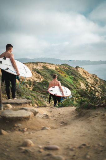 A couple of surfers walking downhill to the coast on a bright cloudy day