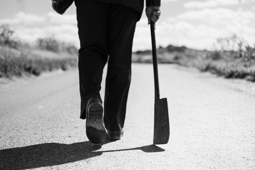 A grayscale shot of a man's legs with an axe on the road