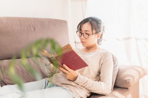 A young Asian woman lying on the couch and reading a book during Covid-19 quarantine