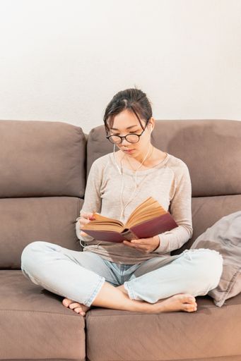 A young Asian woman sitting on the couch and reading a book while listening to music