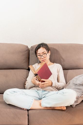 A young Asian woman sitting on the couch and reading a book while listening to music